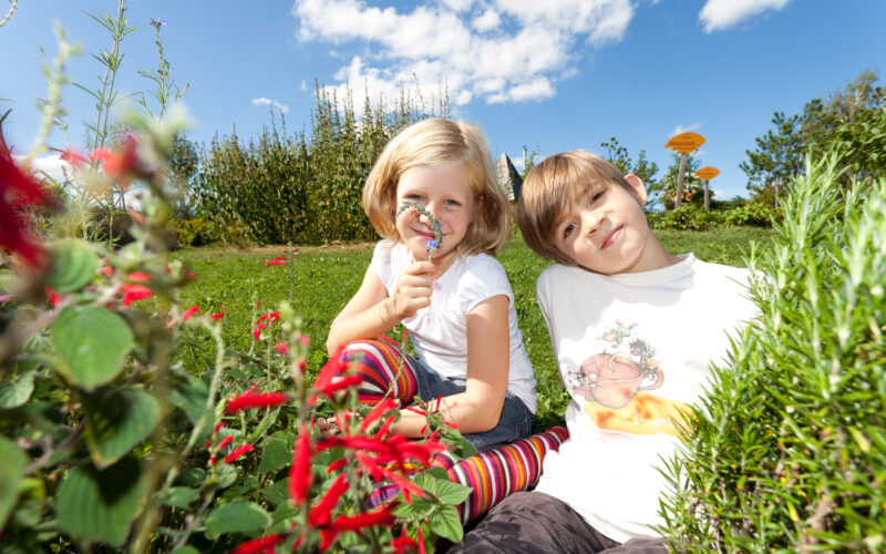 Zwei Kinder sitzen an einem sonnigen Tag im Gras eines Gartens; eines hält sich eine Lupe vors Gesicht, beide sind von roten Blumen und Grün umgeben.