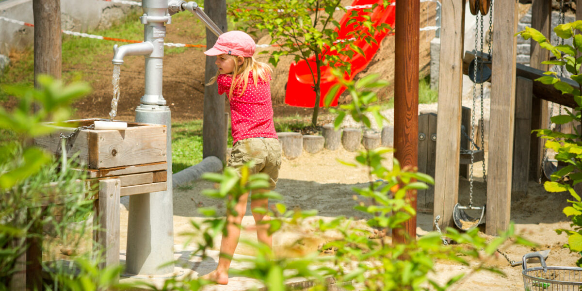 Ein Kind in rosa Hemd und Hut pumpt an einem sonnigen Tag auf einem Spielplatz im Freien Wasser ab, wobei Grünflächen und Spielgeräte zu sehen sind.
