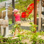 Ein Kind in rosa Hemd und Hut pumpt an einem sonnigen Tag auf einem Spielplatz im Freien Wasser ab, wobei Grünflächen und Spielgeräte zu sehen sind.