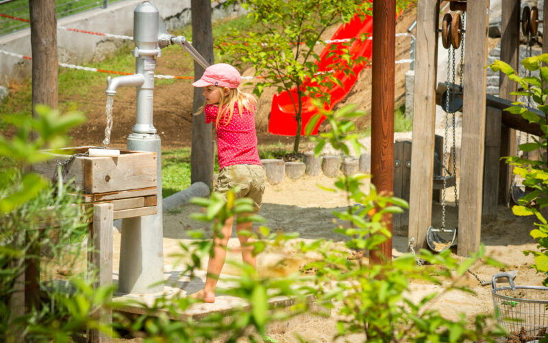 Ein Kind in rosa Hemd und Hut pumpt an einem sonnigen Tag auf einem Spielplatz im Freien Wasser ab, wobei Grünflächen und Spielgeräte zu sehen sind.