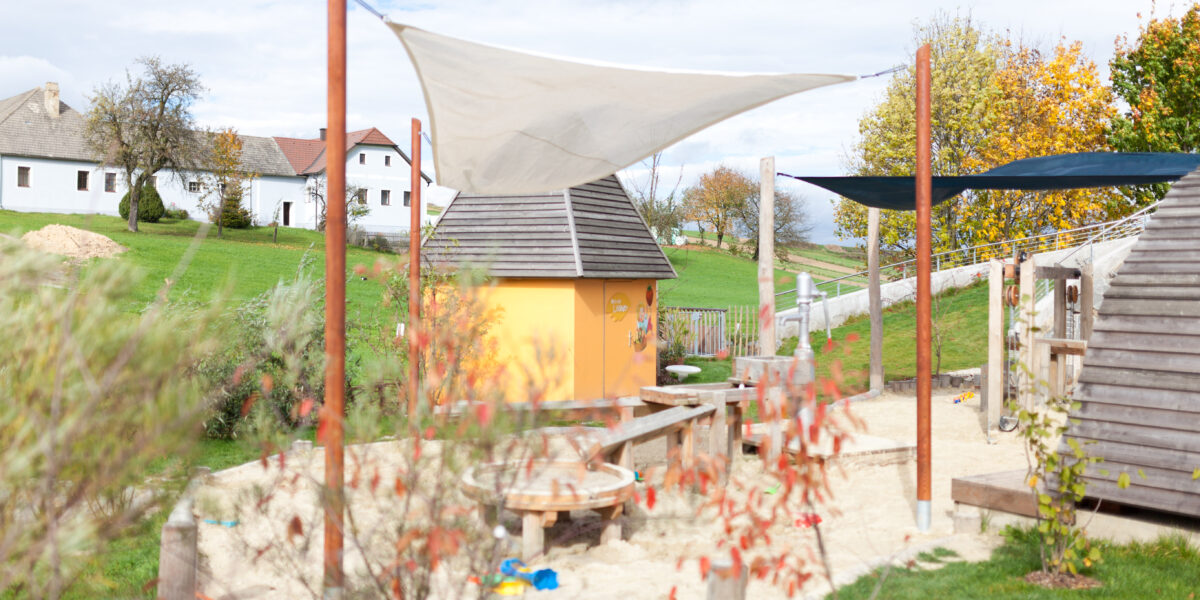 Ein kleiner Spielplatz mit einem Sandkasten, Holzkonstruktionen und einer gelben Hütte, umgeben von Gras, Bäumen und Häusern im Hintergrund.