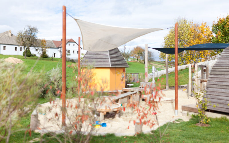 Ein kleiner Spielplatz mit einem Sandkasten, Holzkonstruktionen und einer gelben Hütte, umgeben von Gras, Bäumen und Häusern im Hintergrund.
