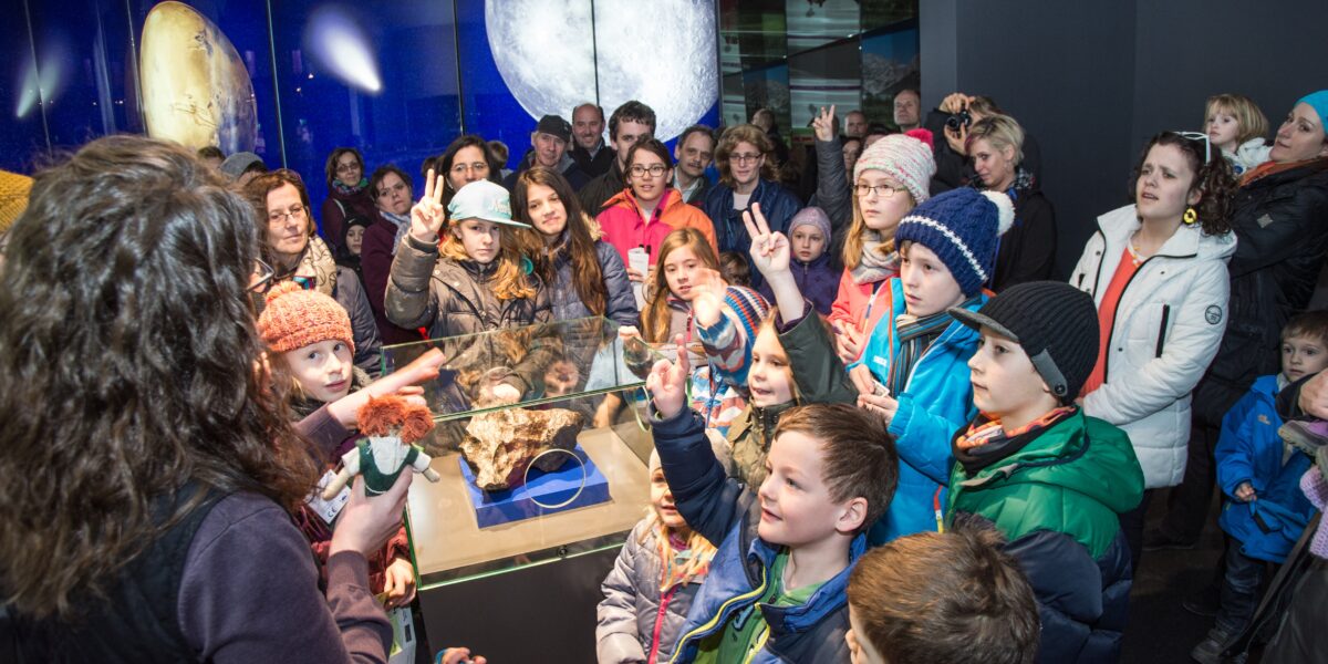 Eine Gruppe von Kindern und Erwachsenen versammelt sich um eine Vitrine in einer Museumsausstellung, während ein Führer ein Objekt zeigt und zur Menge spricht.