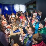 Eine Gruppe von Kindern und Erwachsenen versammelt sich um eine Vitrine in einer Museumsausstellung, während ein Führer ein Objekt zeigt und zur Menge spricht.