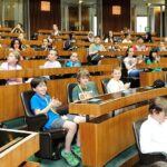 In einem großen Hörsaal sitzen Kinder und Erwachsene in Reihen von gestuften Holzpulten, einige klatschen, andere hören aufmerksam zu.