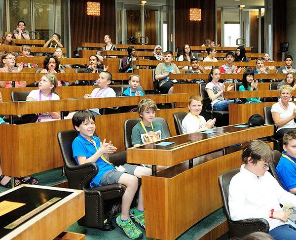 In einem großen Hörsaal sitzen Kinder und Erwachsene in Reihen von gestuften Holzpulten, einige klatschen, andere hören aufmerksam zu.