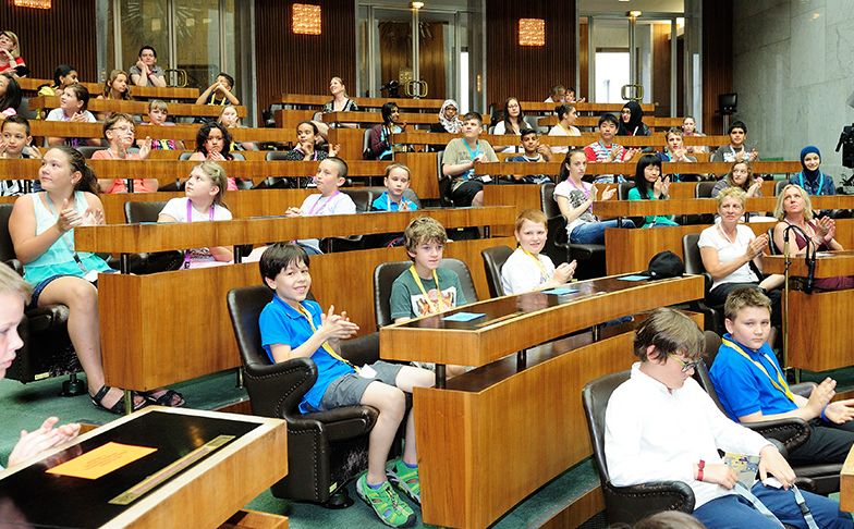 In einem großen Hörsaal sitzen Kinder und Erwachsene in Reihen von gestuften Holzpulten, einige klatschen, andere hören aufmerksam zu.