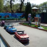 Eine kleine Bahn mit bunten Kinderfahrzeugen, umgeben von Absperrungen und Schildern, in einem Vergnügungspark im Freien.