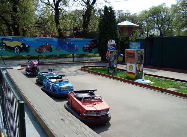 Eine kleine Bahn mit bunten Kinderfahrzeugen, umgeben von Absperrungen und Schildern, in einem Vergnügungspark im Freien.