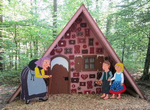 Ein farbenfrohes, dreieckiges Lebkuchenhaus steht in einem Wald, vor dem eine Hexe und zwei Kinder, Hänsel und Gretel, in Form von Comicfiguren stehen.