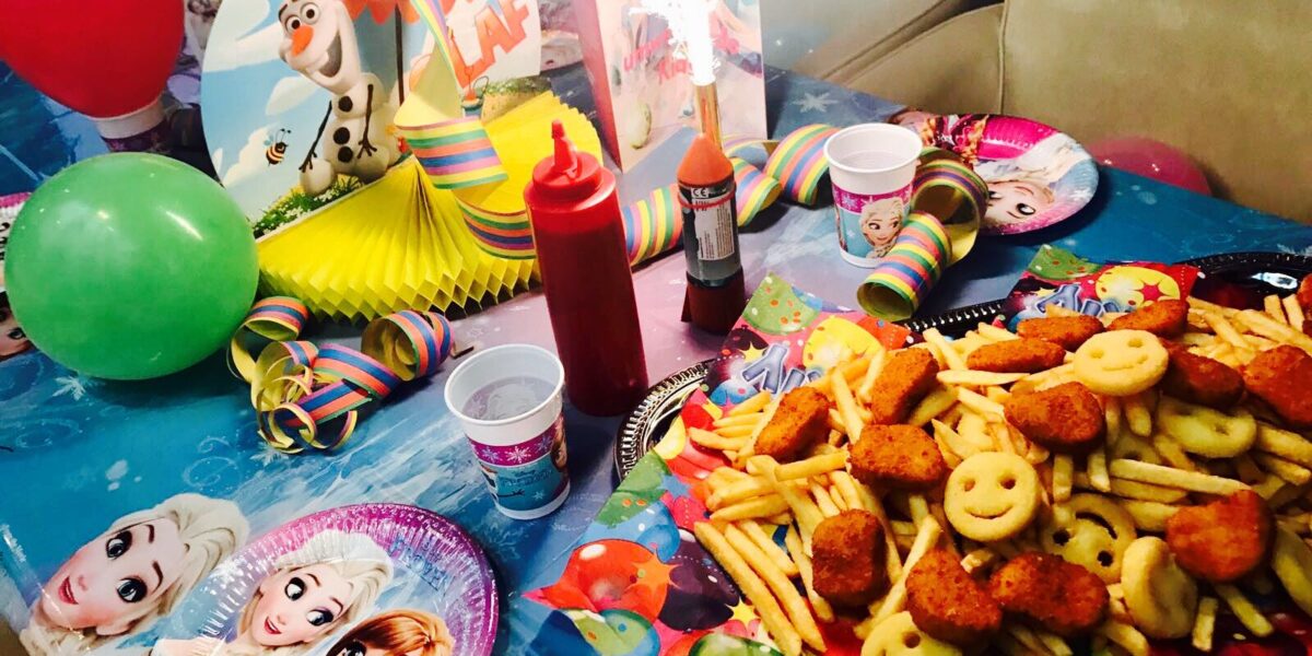 Ein für eine Kinderparty gedeckter Tisch mit Luftballons, bunter Dekoration, Frozen-Tellern und -Bechern, Soßen und einer Platte mit Pommes frites, Smiley-Gesichtern und Nuggets.