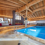 Hallenbad mit klarem, blauem Wasser, Holzbalken an der Decke, Steindetails, umlaufende Sitzgelegenheiten mit roten Kissen und große Fenster, die natürliches Licht hereinlassen.