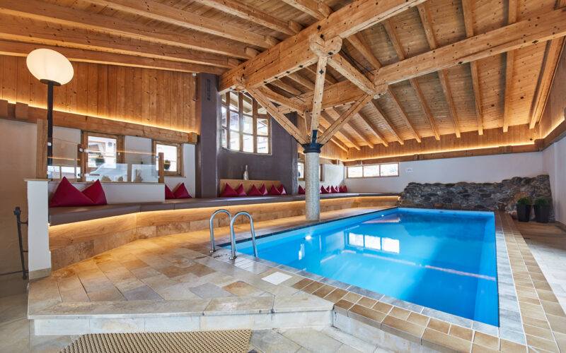 Hallenbad mit klarem, blauem Wasser, Holzbalken an der Decke, Steindetails, umlaufende Sitzgelegenheiten mit roten Kissen und große Fenster, die natürliches Licht hereinlassen.