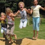 Vier Kinder springen an einem sonnigen Tag im Freien in einen Sandkasten, mit Gras und Bäumen im Hintergrund.