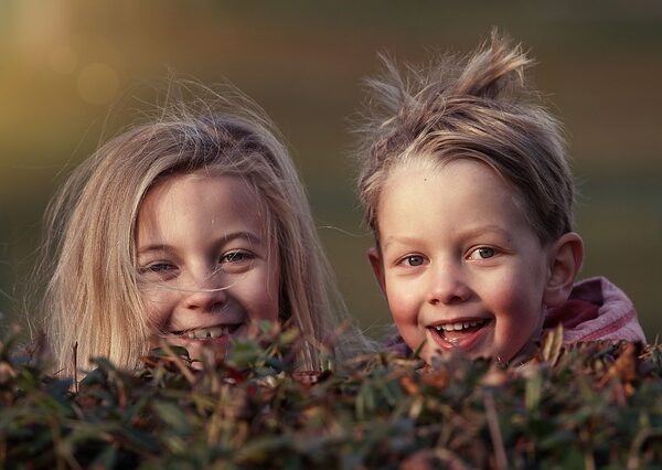 Zwei kleine Kinder mit zerzaustem Haar lächeln in die Kamera, während sie im Freien über einen Busch schauen.