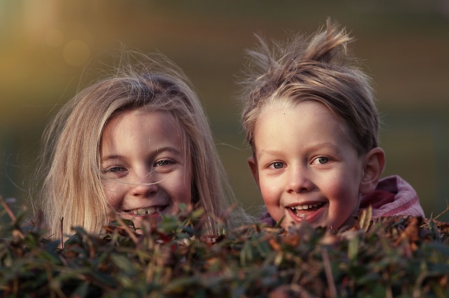 Zwei kleine Kinder mit zerzaustem Haar lächeln in die Kamera, während sie im Freien über einen Busch schauen.