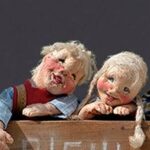 Vier handgefertigte Puppen mit ausdrucksstarken Gesichtern und rosigen Wangen lehnen an einem Holzzaun, jede mit einer einzigartigen Pose und Kleidung vor einem schlichten Hintergrund.