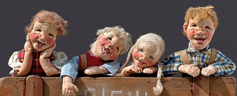 Vier handgefertigte Puppen mit ausdrucksstarken Gesichtern und rosigen Wangen lehnen an einem Holzzaun, jede mit einer einzigartigen Pose und Kleidung vor einem schlichten Hintergrund.