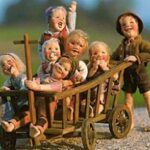 Sechs lächelnde Puppen sitzen in einer hölzernen Schubkarre, während eine siebte Puppe im Freien auf einer Kiesfläche mit unscharfem grünen Hintergrund steht.