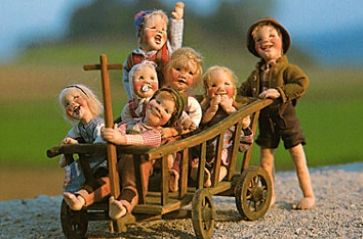 Sechs lächelnde Puppen sitzen in einer hölzernen Schubkarre, während eine siebte Puppe im Freien auf einer Kiesfläche mit unscharfem grünen Hintergrund steht.