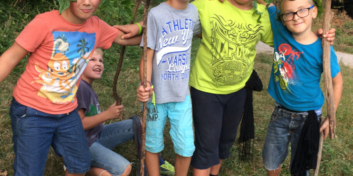 Vier Jungen stehen im Freien zusammen, halten Stöcke in der Hand und tragen Stirnbänder mit Blättern, während ein weiteres Kind im Hintergrund sitzt. Die Szene spielt in einer grasbewachsenen, bewaldeten Gegend. Lucy Lynn-Logo in der unteren rechten Ecke.