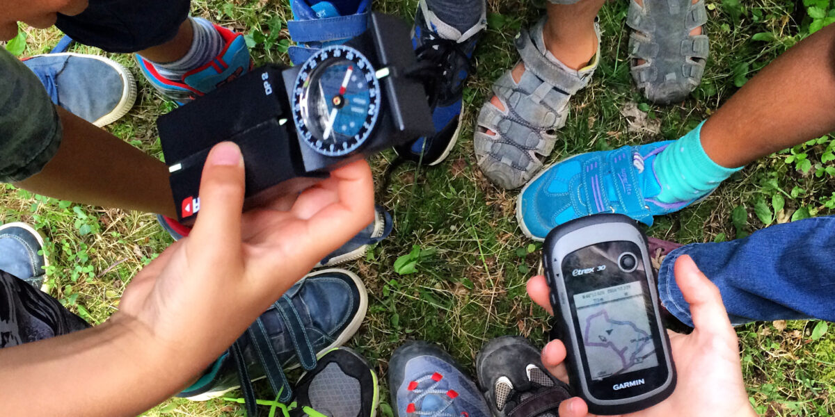 Eine Gruppe von Kindern steht im Freien in einem Kreis, einer hält einen Kompass, ein anderer ein GPS-Gerät, die Füße stehen dicht beieinander im Gras.