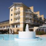 Ein modernes, gelbes Hotelgebäude mit mehreren Balkonen überblickt einen Außenpool mit einem zentralen Springbrunnen unter einem klaren blauen Himmel.