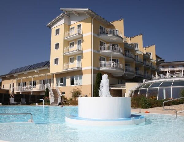 Ein modernes, gelbes Hotelgebäude mit mehreren Balkonen überblickt einen Außenpool mit einem zentralen Springbrunnen unter einem klaren blauen Himmel.