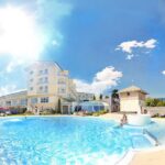 Ein heller, sonniger Tag an einem Hotel-Freibad mit klarem, blauem Wasser, einer schwimmenden Person und dem Hotelgebäude im Hintergrund.