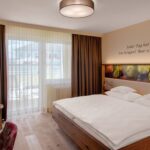 Ein Hotelzimmer mit einem Doppelbett, Holzmöbeln, einem Schreibtisch mit Stühlen, einem großen Fenster mit durchsichtigen Vorhängen und einem Zitat in deutscher Sprache an der Wand über dem Bett.