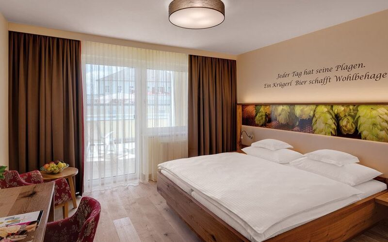 Ein Hotelzimmer mit einem Doppelbett, Holzmöbeln, einem Schreibtisch mit Stühlen, einem großen Fenster mit durchsichtigen Vorhängen und einem Zitat in deutscher Sprache an der Wand über dem Bett.