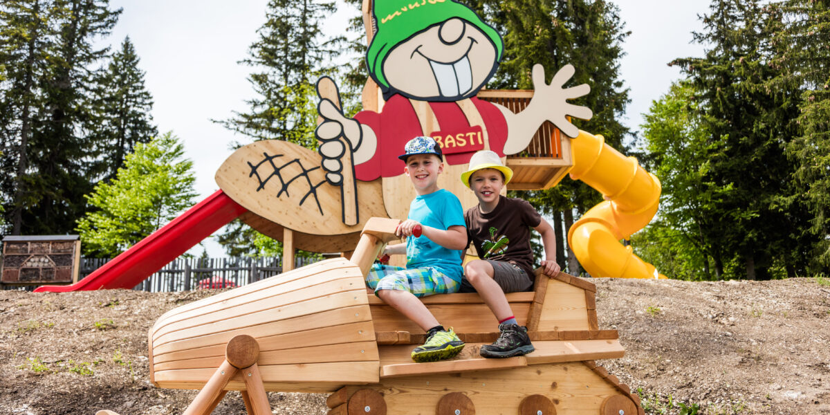Zwei Kinder sitzen auf einem hölzernen Spielgerät in Form eines Schneemobils, mit einem großen Cartoon-Zwerg und Rutschen im Hintergrund auf einem Spielplatz im Freien.