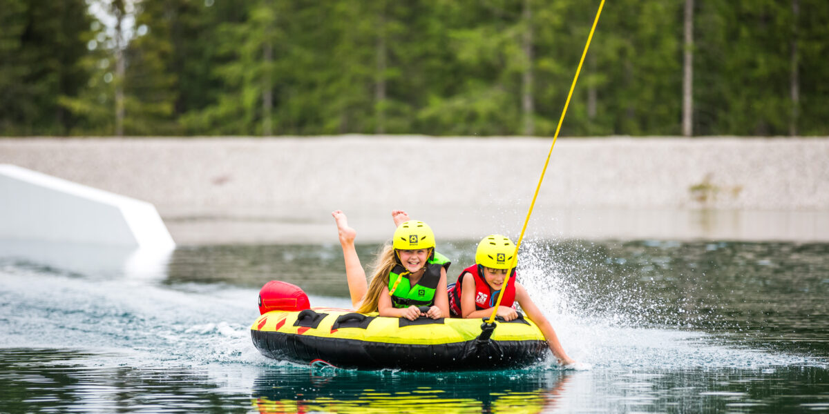 Zwei Personen mit Helmen und Schwimmwesten fahren auf einem aufblasbaren Schlauch, der von einem Seil gezogen wird, auf einem See, mit plätscherndem Wasser und Bäumen im Hintergrund.