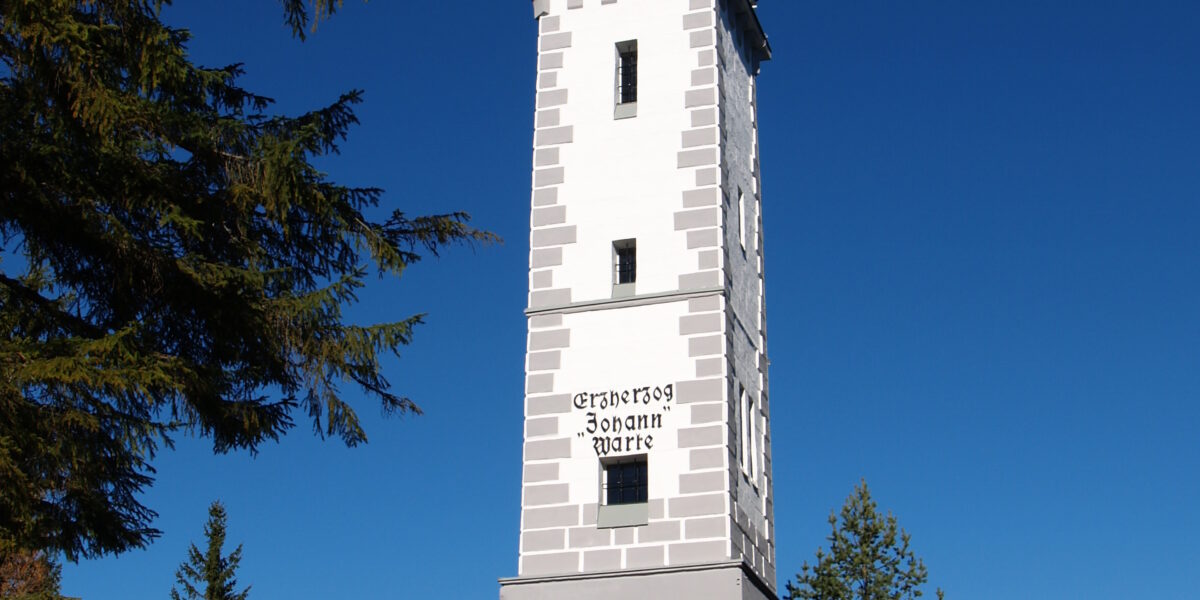 Ein hoher, weißer steinerner Aussichtsturm mit spitzem Dach steht auf einem grasbewachsenen Hügel, umgeben von immergrünen Bäumen unter einem klaren blauen Himmel.