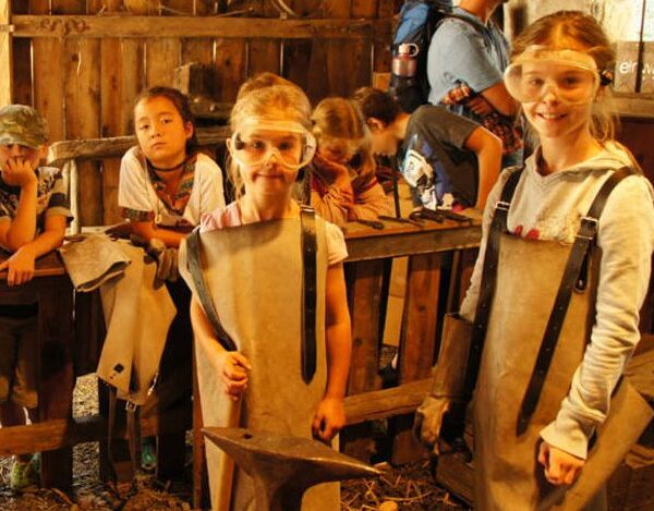 Kinder mit Schutzbrillen und Schürzen stehen in einer Werkstatt mit Schmiedewerkzeugen, einige lächeln, in einem scheunenähnlichen Holzgebäude.