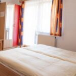 Ein einfaches, aufgeräumtes Schlafzimmer mit einem Doppelbett, hellen Holzmöbeln, einem an der Wand montierten Fernseher, einem Spiegel, Kleiderhaken und bunten Vorhängen an einem Fenster.