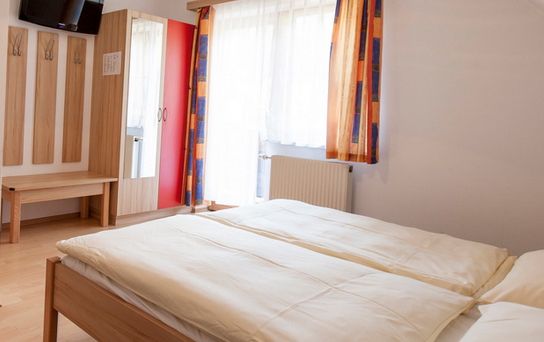 Ein einfaches, aufgeräumtes Schlafzimmer mit einem Doppelbett, hellen Holzmöbeln, einem an der Wand montierten Fernseher, einem Spiegel, Kleiderhaken und bunten Vorhängen an einem Fenster.