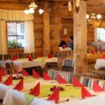 Ein rustikales Restaurant mit Wänden und Möbeln aus Holzbalken, ordentlich gedeckten Tischen mit weißen Tüchern und roten gefalteten Servietten und warmer Beleuchtung.