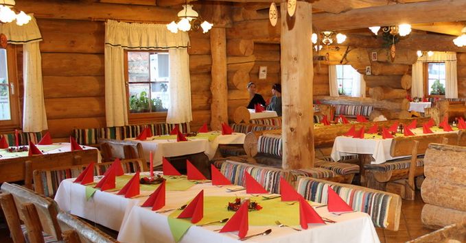 Ein rustikales Restaurant mit Wänden und Möbeln aus Holzbalken, ordentlich gedeckten Tischen mit weißen Tüchern und roten gefalteten Servietten und warmer Beleuchtung.