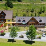 Zwei große Holzhütten mit Balkonen und einer roten Spielplatzrutsche stehen auf einem grasbewachsenen Hügel, mit mehreren geparkten Autos und Bäumen im Vordergrund.