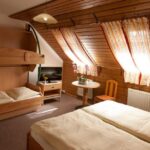 Ein gemütliches Zimmer im Dachgeschoss mit Holzwänden, einem Doppelbett, Etagenbetten, einem kleinen Tisch mit zwei Stühlen und verdunkelten Fenstern, die Tageslicht hereinlassen.