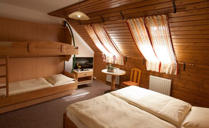 Ein gemütliches Zimmer im Dachgeschoss mit Holzwänden, einem Doppelbett, Etagenbetten, einem kleinen Tisch mit zwei Stühlen und verdunkelten Fenstern, die Tageslicht hereinlassen.