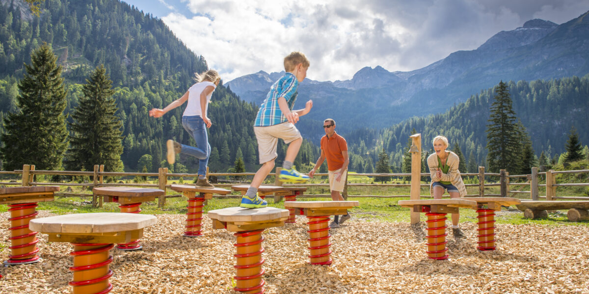 Vier Personen, darunter zwei Kinder, spielen auf großen runden Plattformen mit roten Federn auf einem Spielplatz im Freien, der von Bergen und Bäumen umgeben ist.