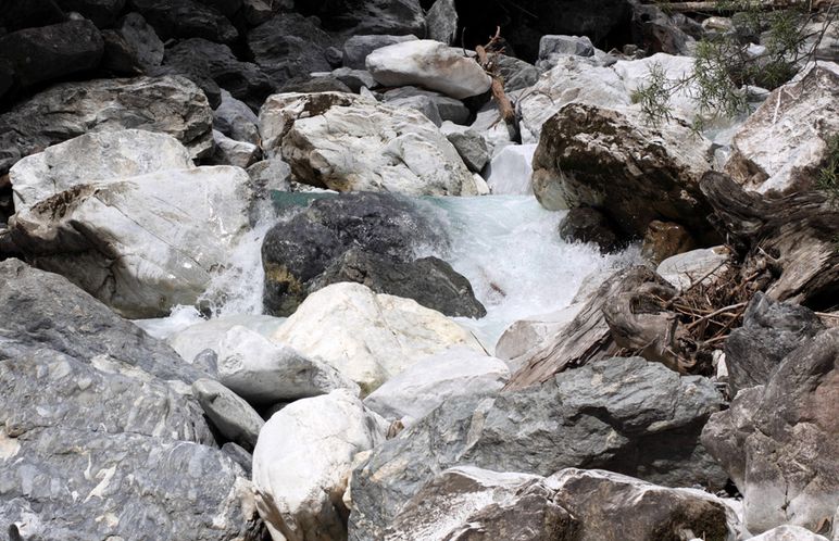 Ein kleiner Wasserlauf fließt über und zwischen großen, unregelmäßigen Felsen und Blöcken, die von einigen Baumzweigen umgeben sind.