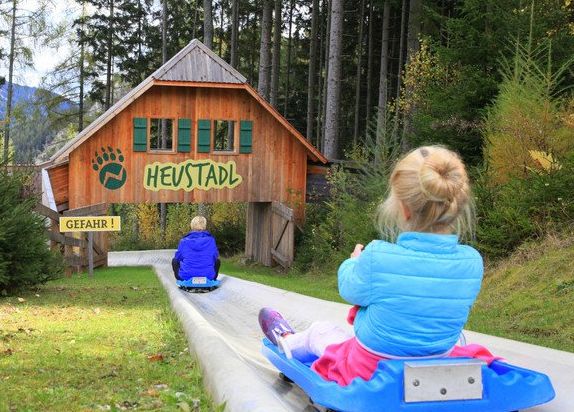 Zwei Kinder fahren auf blauen Schlitten eine Betonrutsche hinunter zu einer Holzhütte mit der Aufschrift "HEUSTADL" in einem Waldgebiet. Links ist ein gelbes Warnschild "GEFAHR!" zu sehen.
