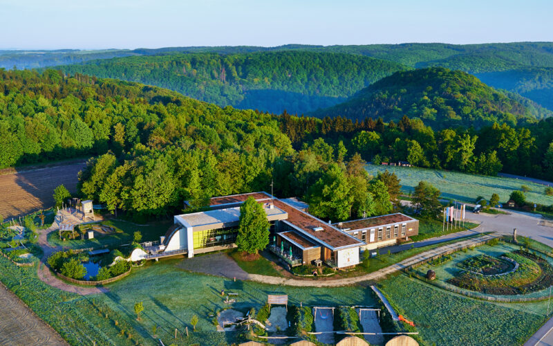 Luftaufnahme eines modernen Gebäudes, umgeben von Bäumen und Hügeln, mit Gärten, Wegen und einem Parkplatz im Vordergrund.