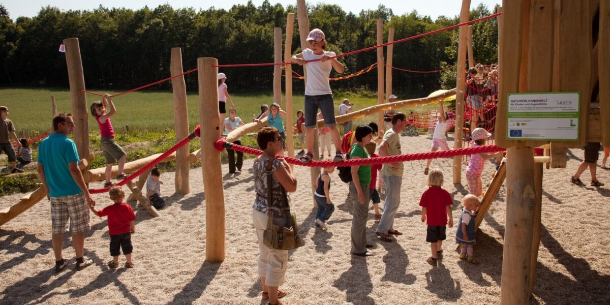 Kinder und Erwachsene treffen sich auf einem Außenspielplatz mit Holzkonstruktionen und roten Seilen, umgeben von Bäumen und einer Wiese.