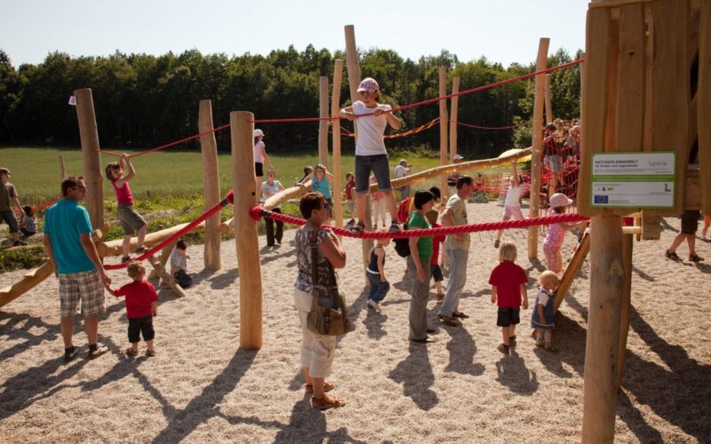 Kinder und Erwachsene treffen sich auf einem Außenspielplatz mit Holzkonstruktionen und roten Seilen, umgeben von Bäumen und einer Wiese.