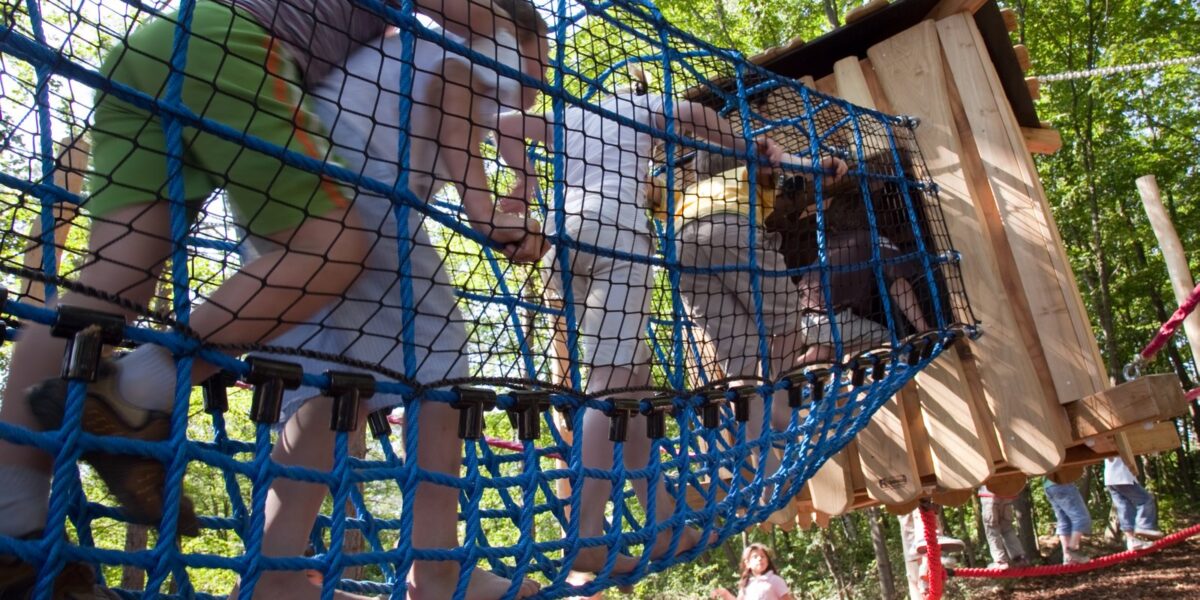 Kinder klettern über eine blaue Seilbrücke auf einem Außenspielplatz mit Bäumen und einer Holzkonstruktion im Hintergrund.