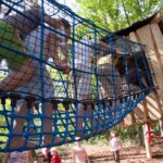 Kinder klettern über eine blaue Seilbrücke auf einem Außenspielplatz mit Bäumen und einer Holzkonstruktion im Hintergrund.